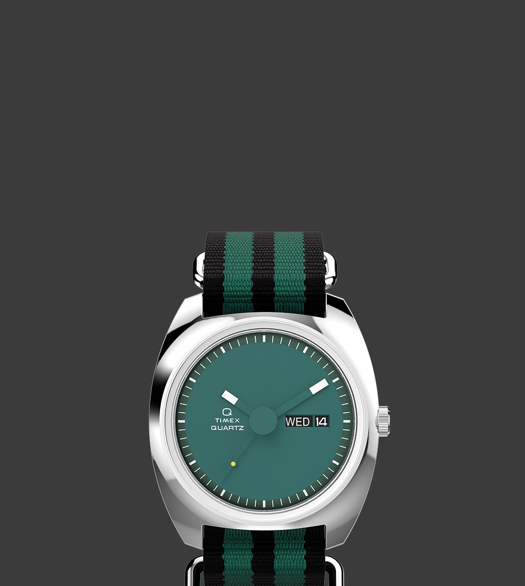 Front view of the Q Timex® 1975 Enigma watch showcasing a deep green dial, bold white hour markers, and a day-date window at 3 o'clock. The watch features a striped green and black fabric strap.