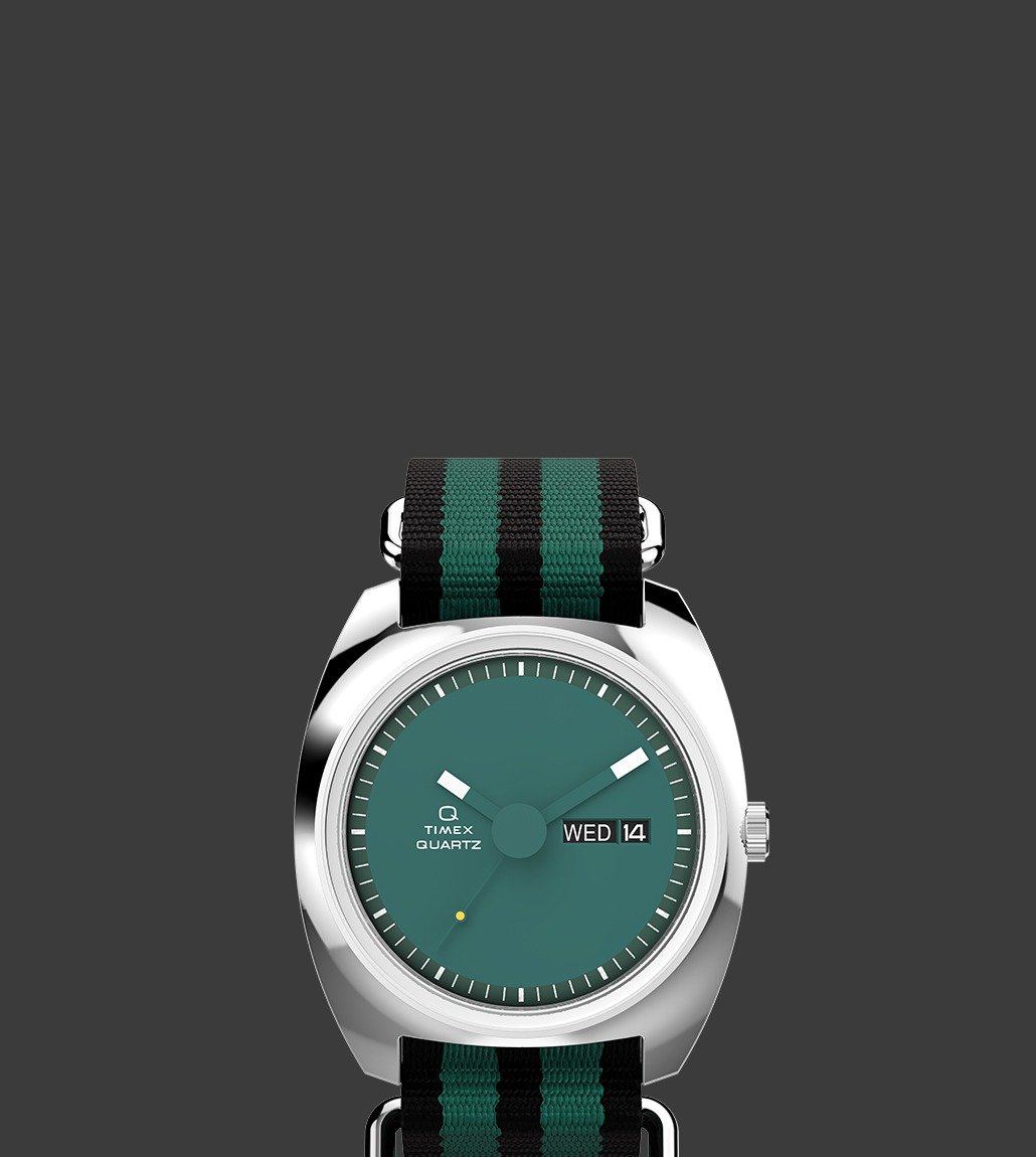 Front view of the Q Timex® 1975 Enigma watch showcasing a deep green dial, bold white hour markers, and a day-date window at 3 o'clock. The watch features a striped green and black fabric strap.