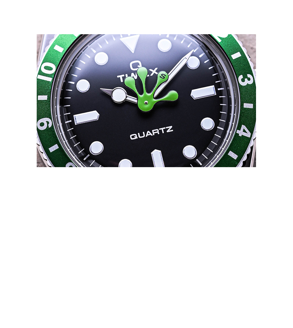 Macro close-up of the Timex x seconde/seconde/ watch dial showcasing the black face with bold white hour markers, silver-tone hands, and a bright green frog-shaped hour hand at center. The green bezel with white numerals frames the dial, emphasizing the quirky artistic detail.