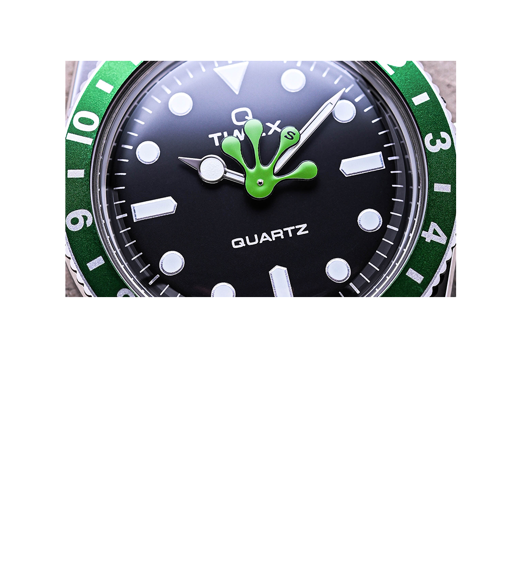 Macro close-up of the Timex x seconde/seconde/ watch dial showcasing the black face with bold white hour markers, silver-tone hands, and a bright green frog-shaped hour hand at center. The green bezel with white numerals frames the dial, emphasizing the quirky artistic detail.