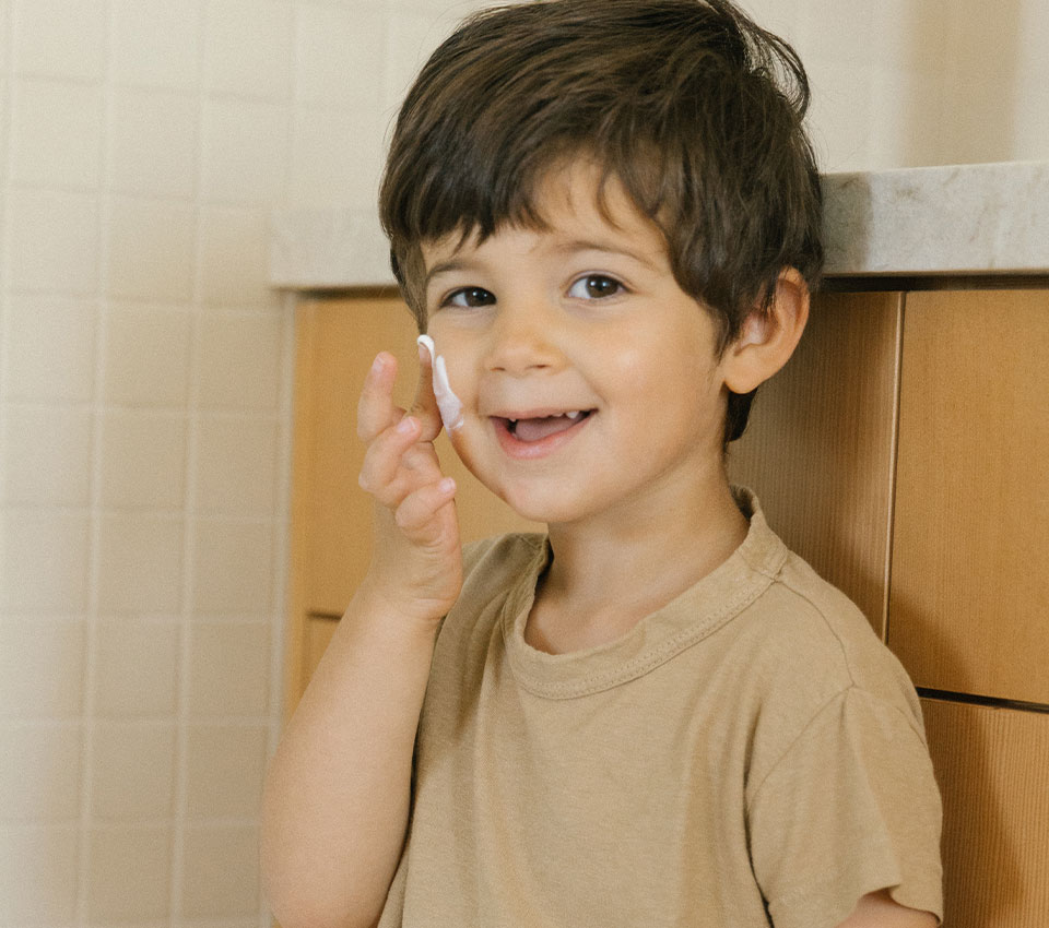Smiling child applying cream to cheek.