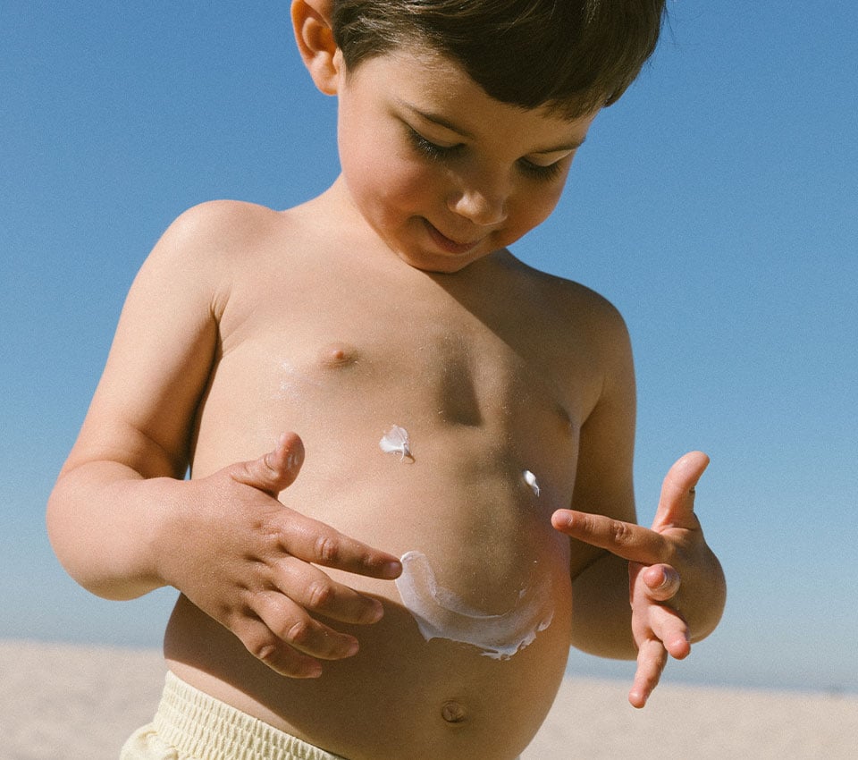 Young boy applying sunscreen to his stomach at the beach.
