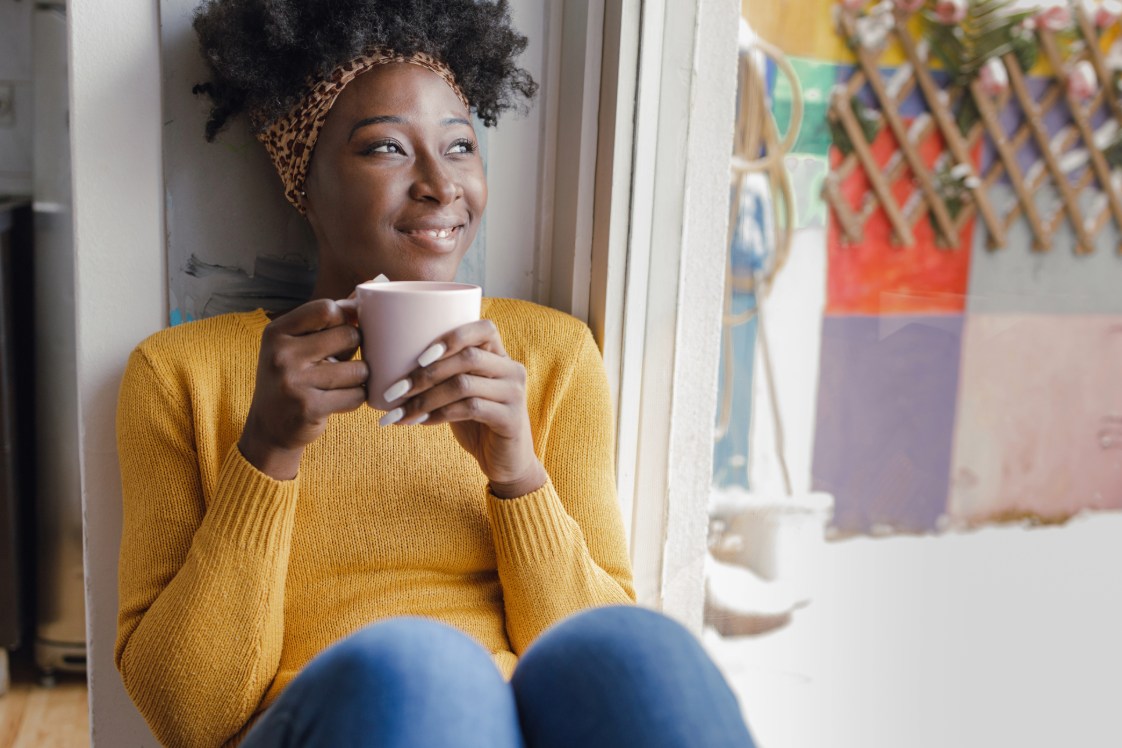 Person in a yellow top holding a cup while looking out the window.