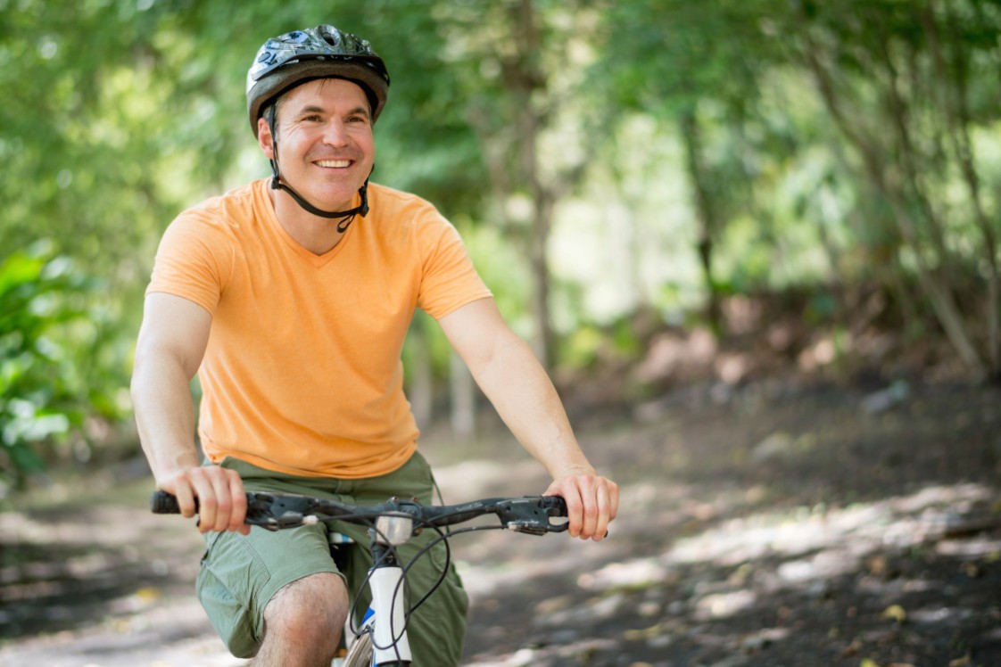 Adult riding a mountain bike on a forest trail, wearing a helmet and casual outdoor clothing.