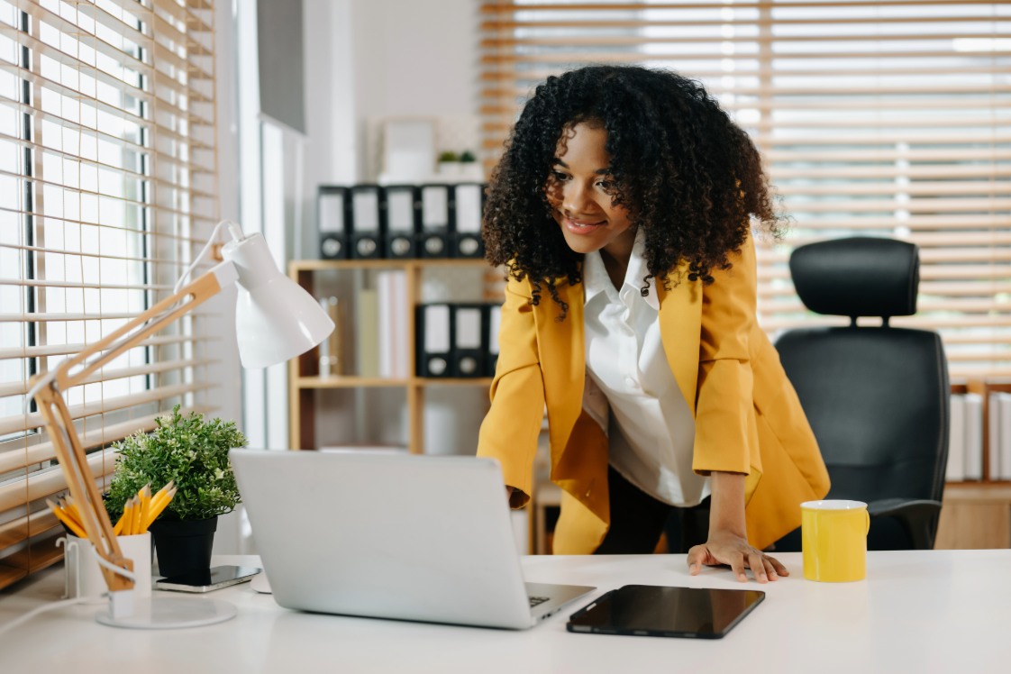 An adult in a yellow blazer in office environment on their laptop.