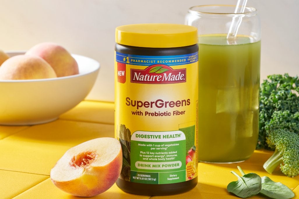 Nature Made SuperGreens with Prebiotic Fiber tub with a glass of green liquid, green vegetables on a yellow counter.