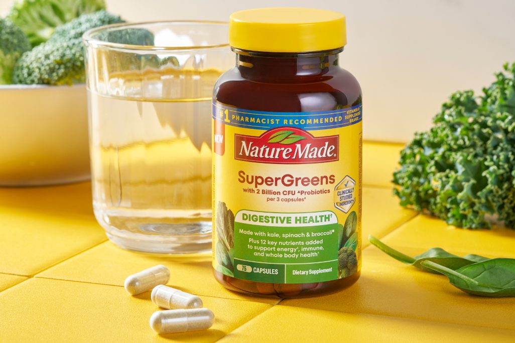 Nature Made SuperGreens with Probiotics Capsules bottle with a class of water and green vegetables on a yellow kitchen counter.