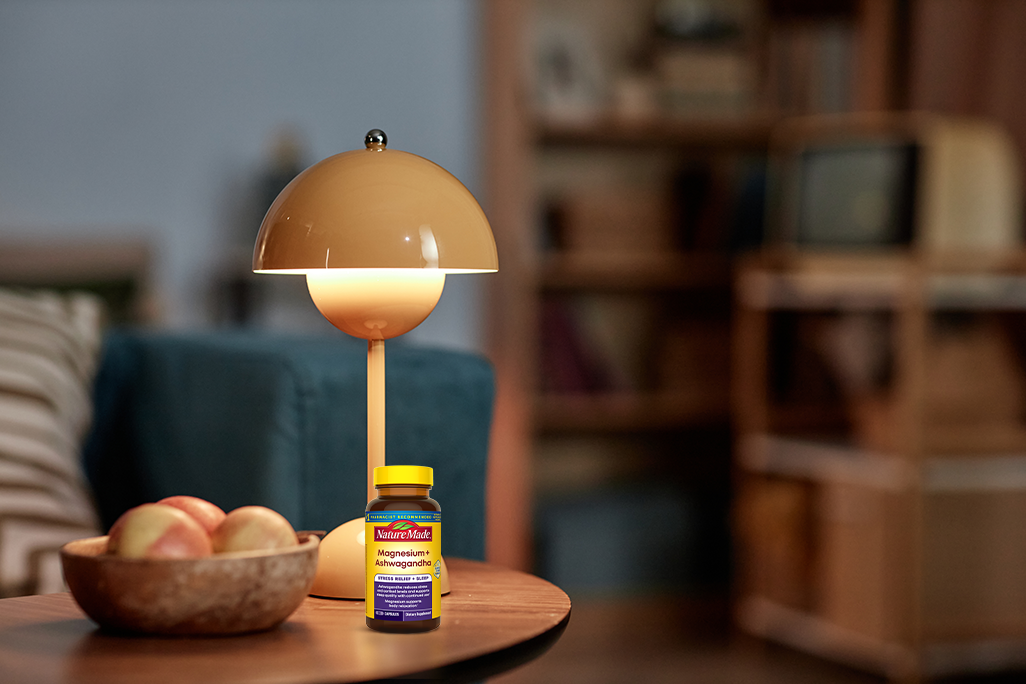 Nature Made Magnesium + Ashwagandha Capsules on a side table with a lamp in a cozy living room.