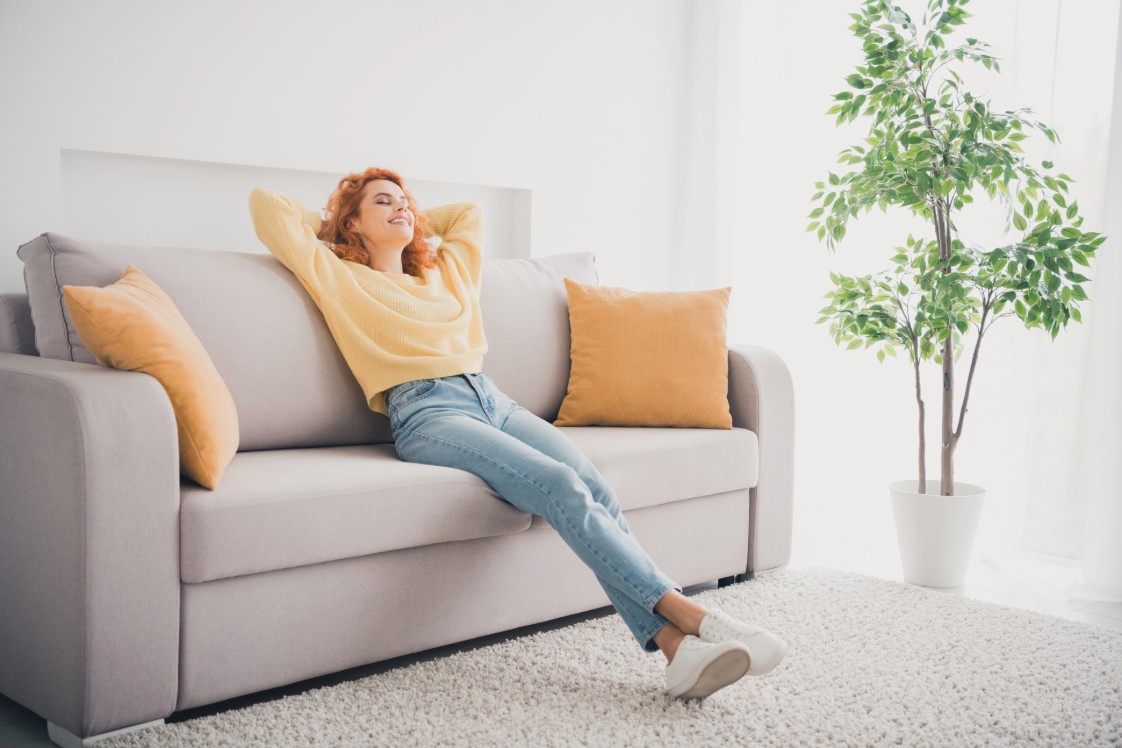 A person in a yellow sweater and blue jeans leaning back on the couch with throw pillows and a plant in the background. 
