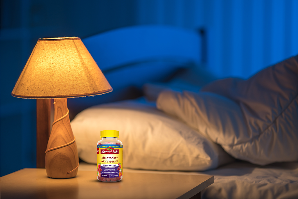Nature Made Melatonin + Magnesium Sleep and Relax Gummies on a bedside table with a lamp and bed at night.