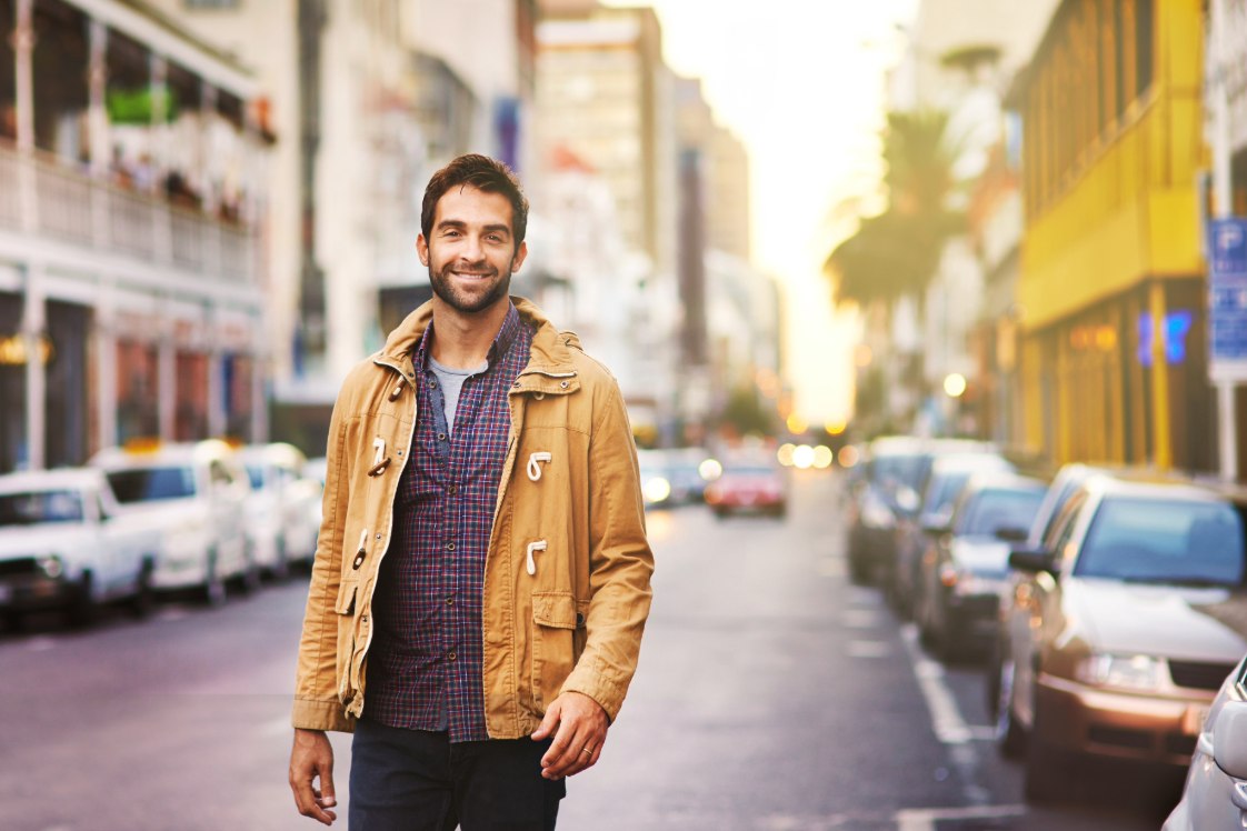 Man walking down a city street at sunset with parked cars and urban buildings.