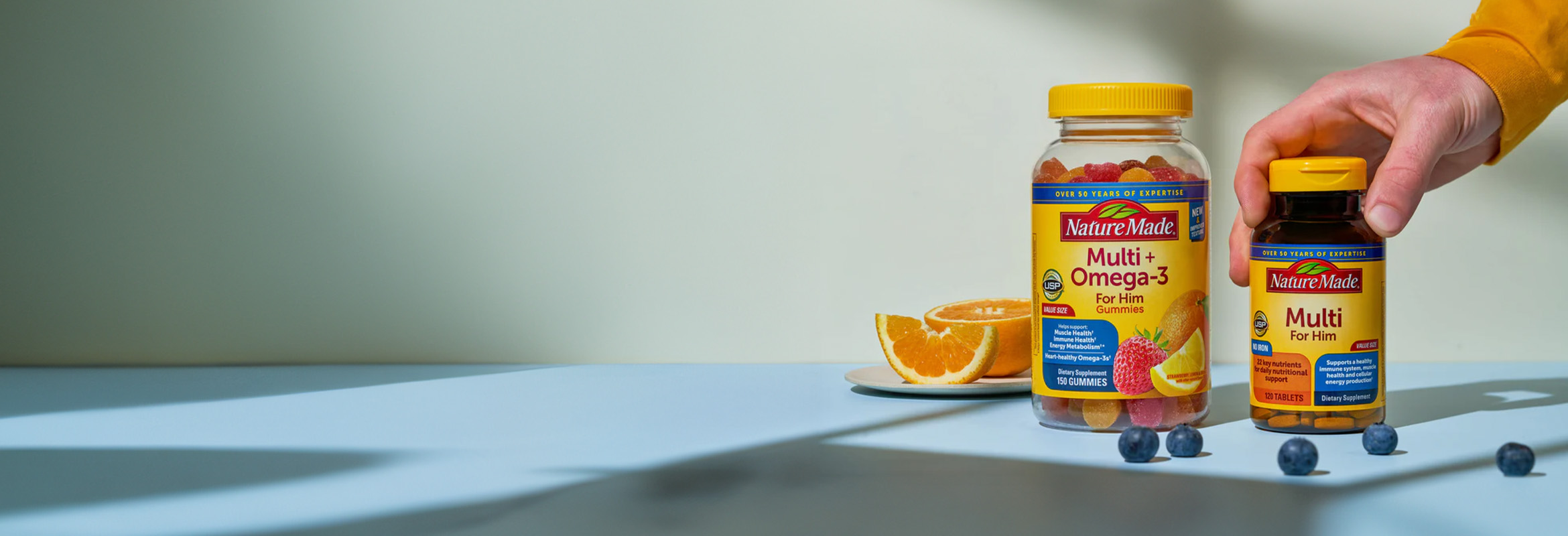 Nature Made Multi + Omega‑3 and men’s multivitamin supplements displayed with fresh fruit on a tabletop.