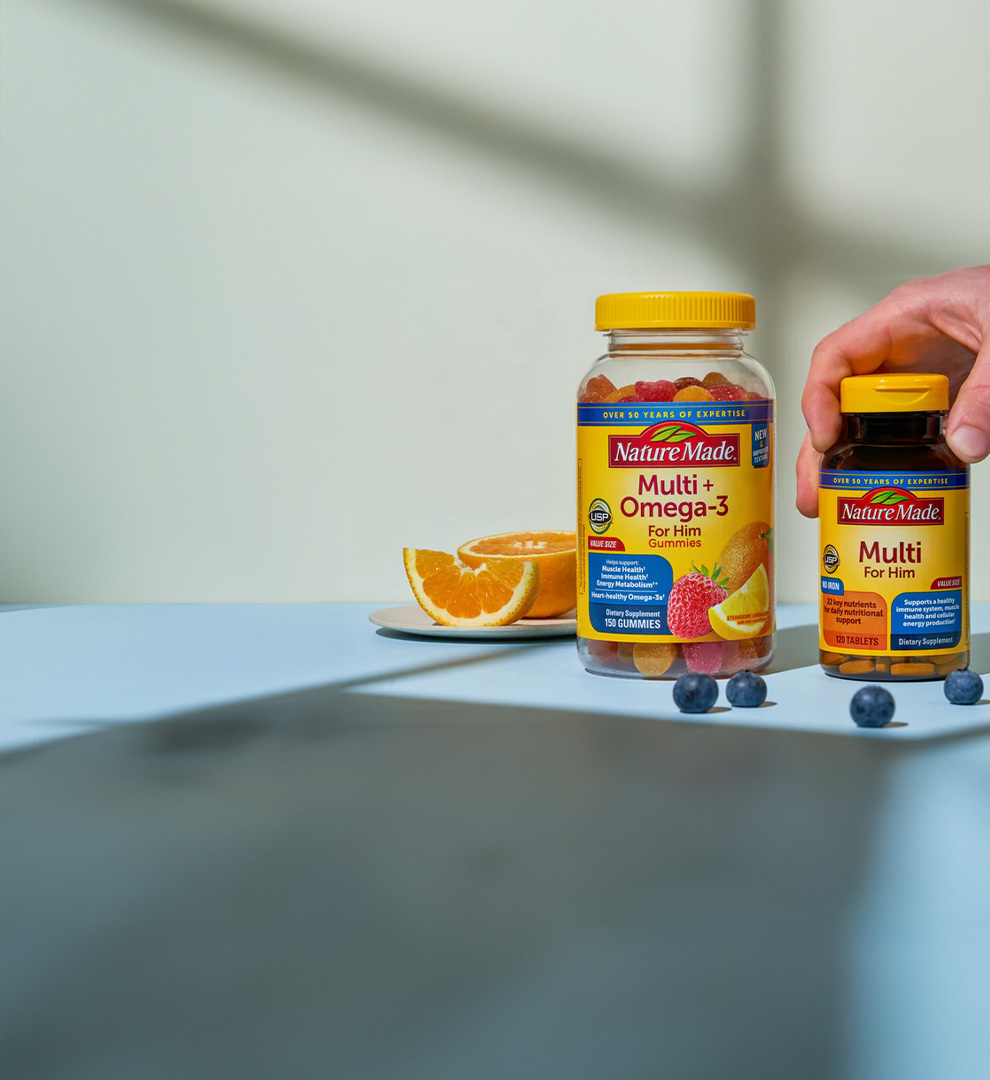Nature Made Multi + Omega‑3 and men’s multivitamin supplements displayed with fresh fruit on a tabletop.