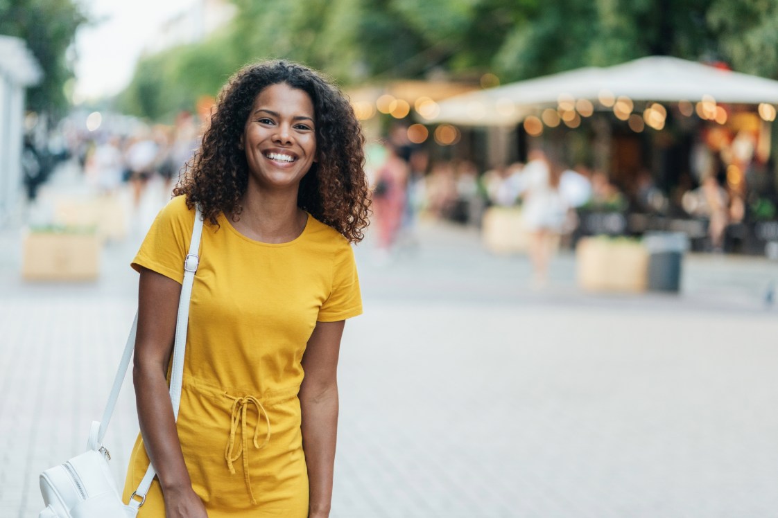 Woman in a yellow dress walking through a lively outdoor city street with cafés and pedestrians.