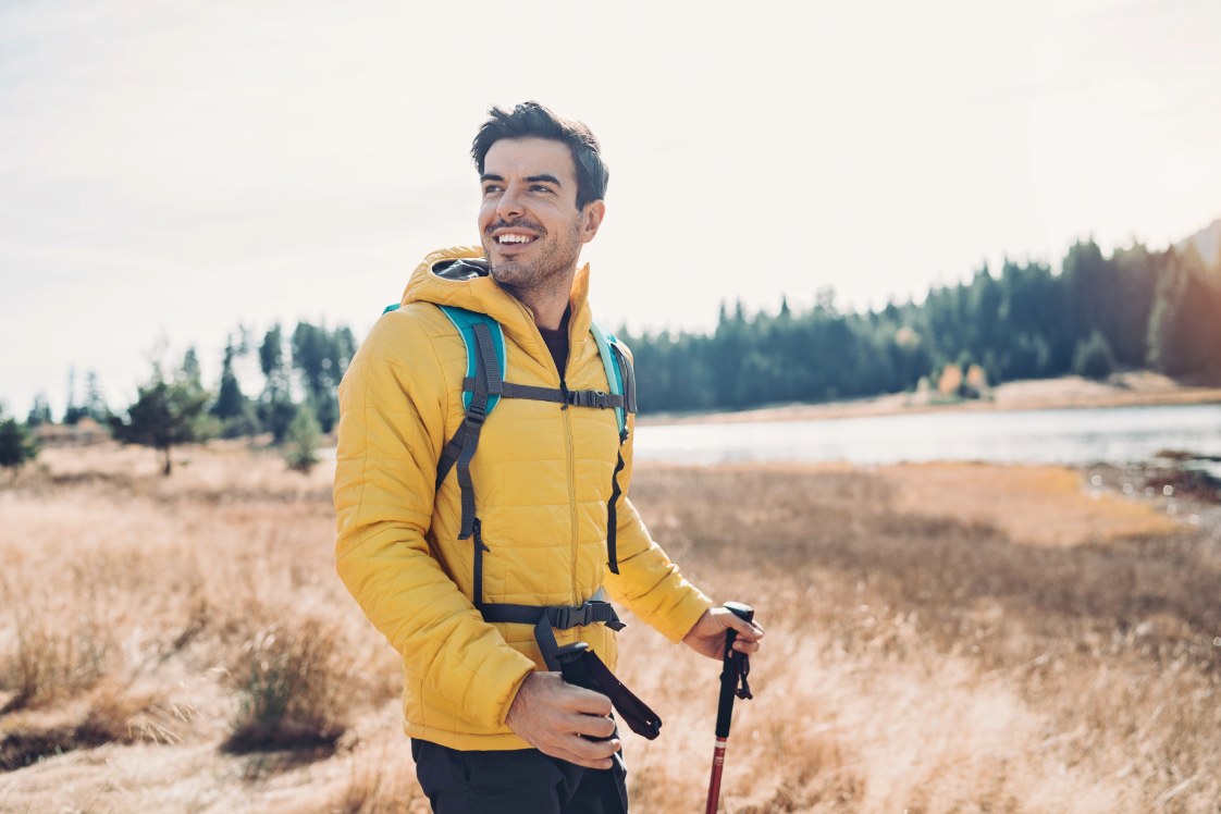 Adult in a yellow jacket smiling outdoors with trees in the background.
