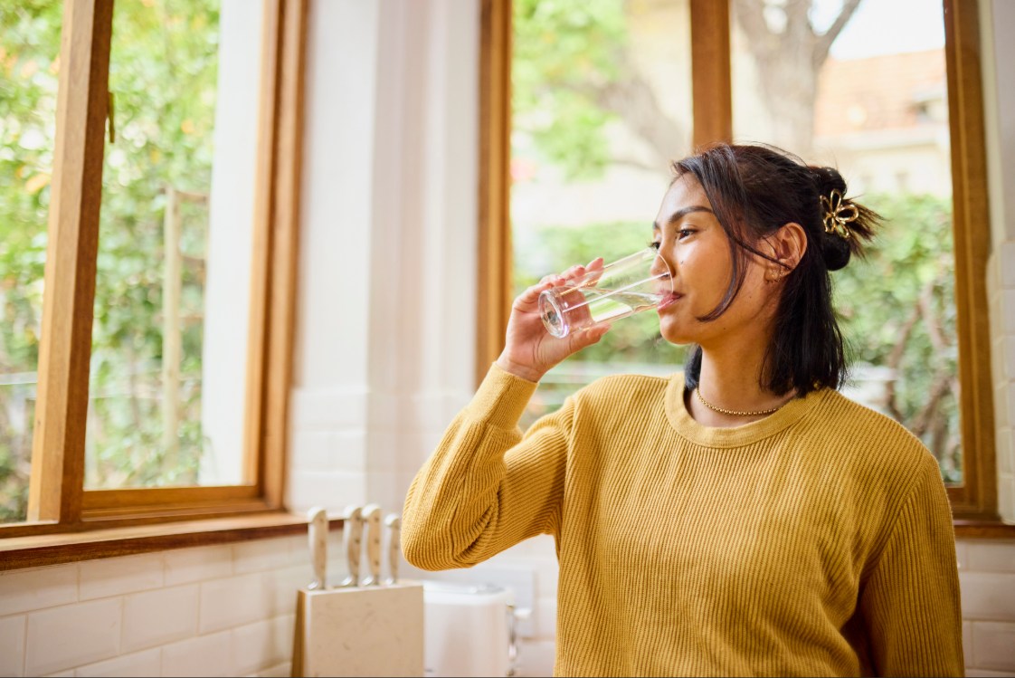 Adult in yellow sweater drinking water looking out of a window. 