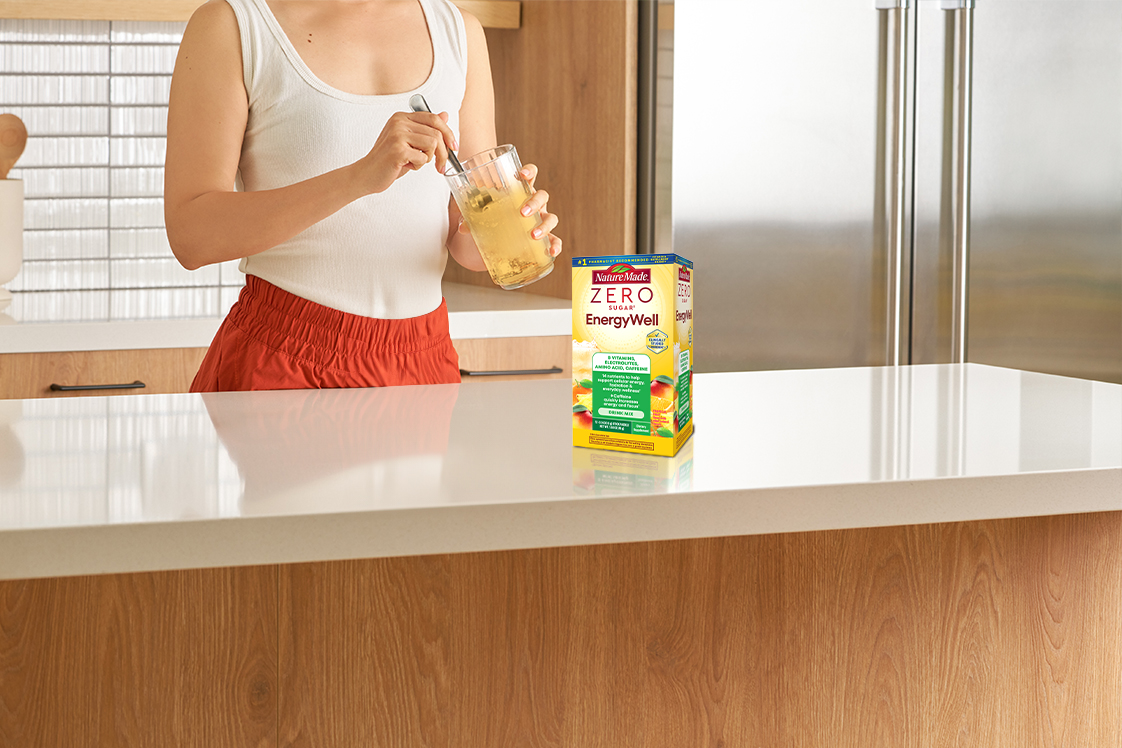 Nature Made Zero Sugar EnergyWell Drink Mix on a kitchen counter with adult stirring a glass of water.
