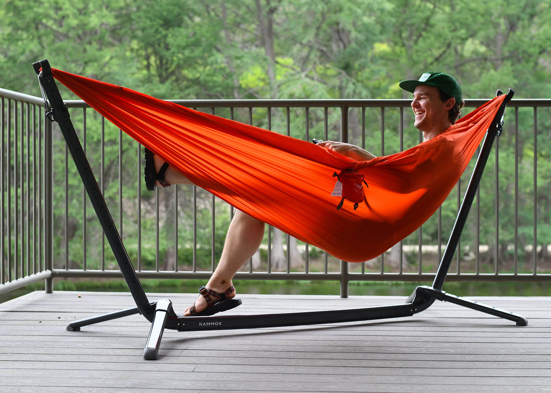 Man sits in ember orange Roo Single hammock hung from Swiftlet portable hammock stand on a patio deck over water.