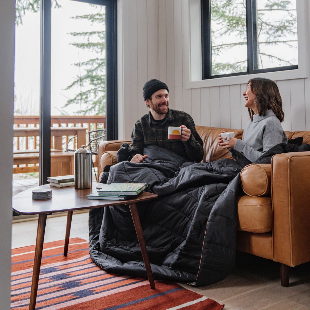 Two people cozying up on a couch with a Rumpl 2-person blanket and mugs of hot beverages.