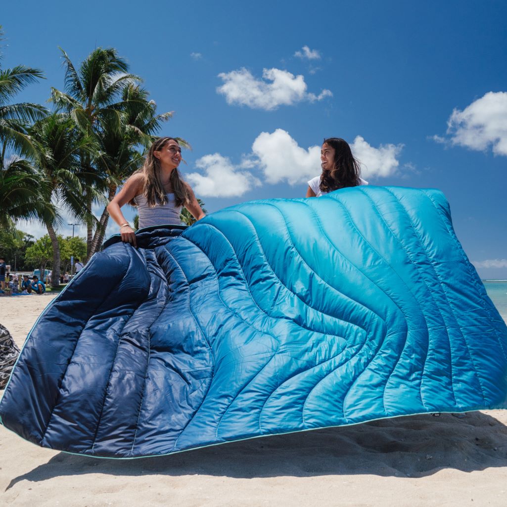 Two girls dusting the sand off a 2-person sized blanket while on the beach.
