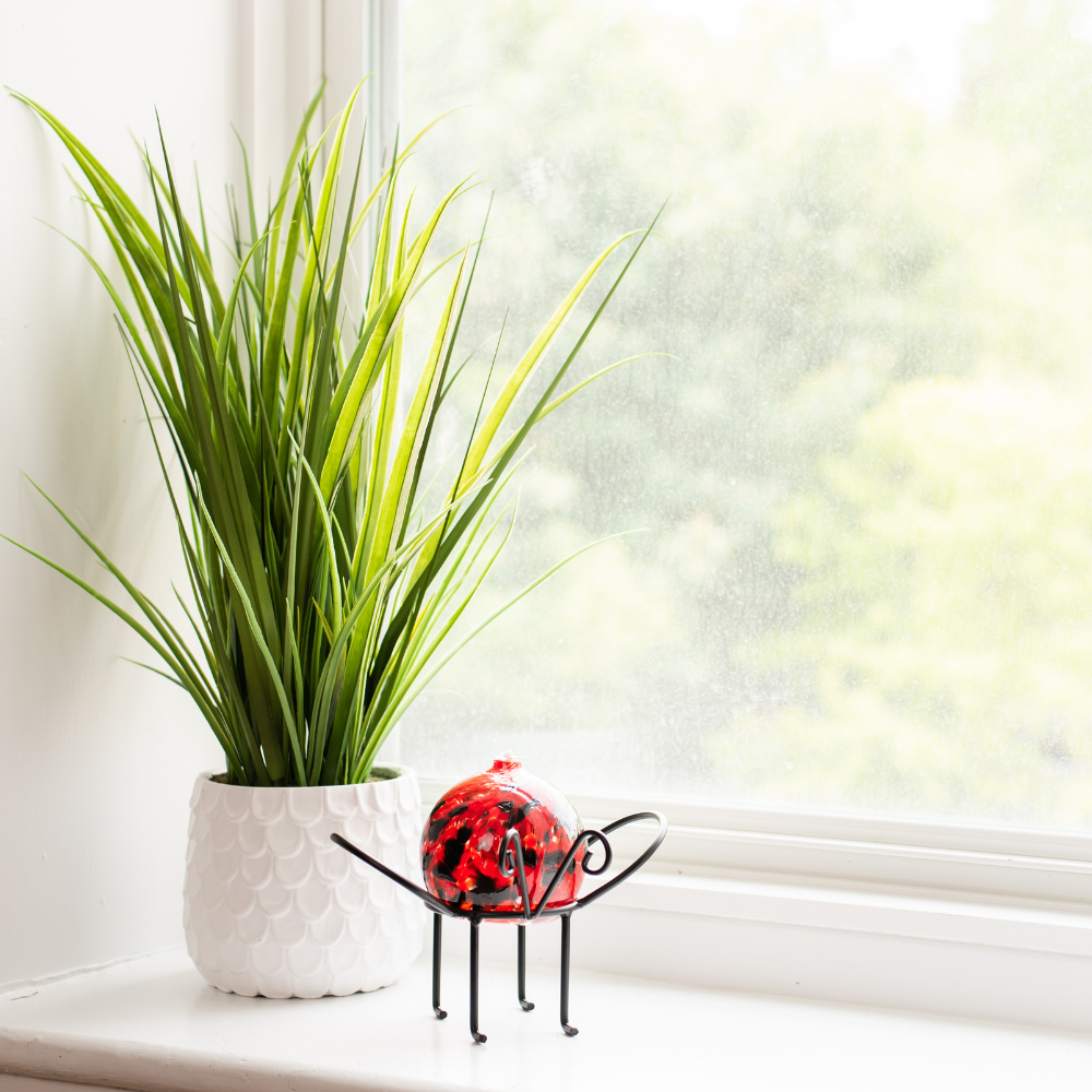 Ladybird Red Nature's Whimsy Oil Lamp on an Insect Garden Holder. Displayed on a white windowsill in front of a plant. 