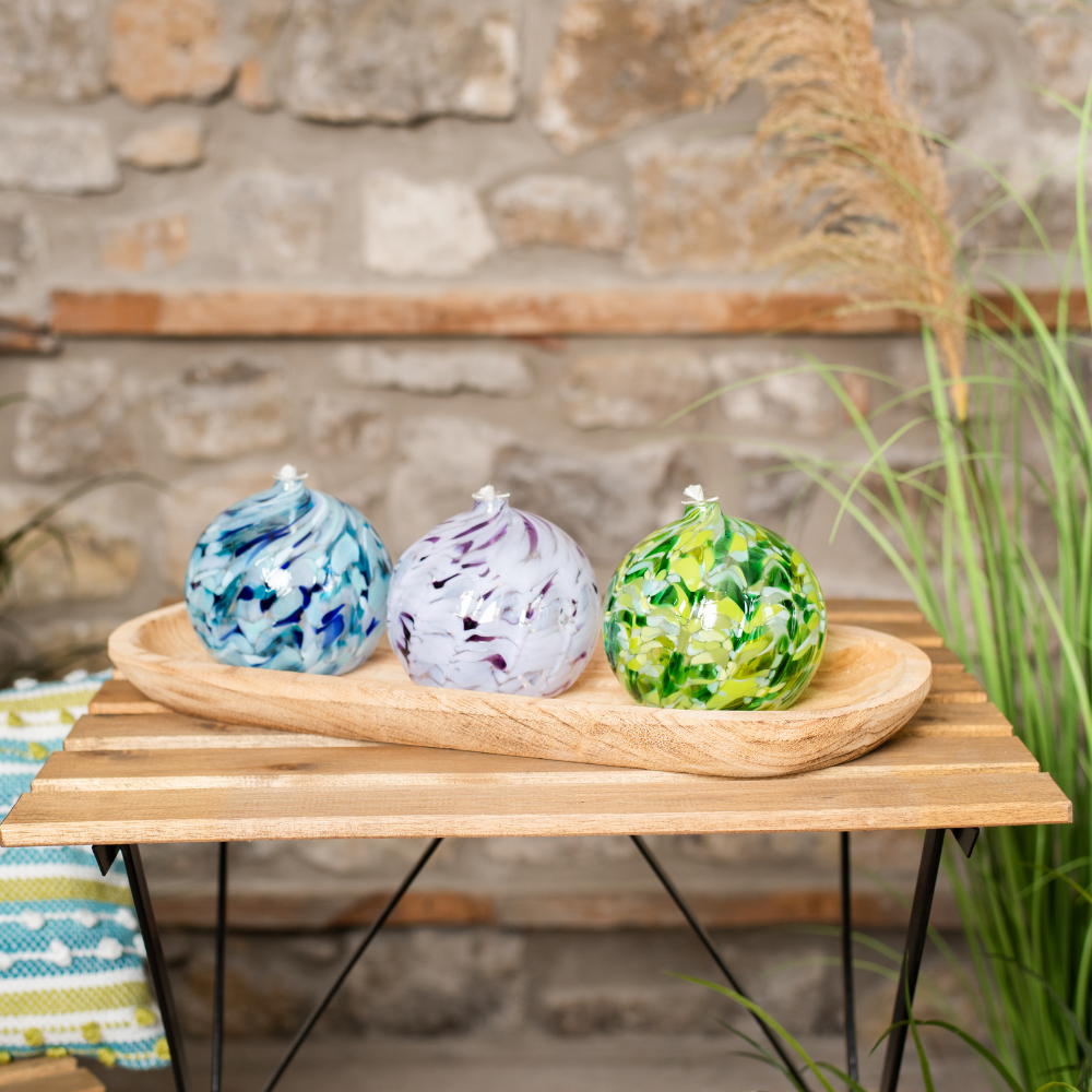 Dark and light blue and white round oil lamp, purple and white round oil lamp and dark and light green and white round oil lamp on a wood tray on a wood table. Stone wall in the background.