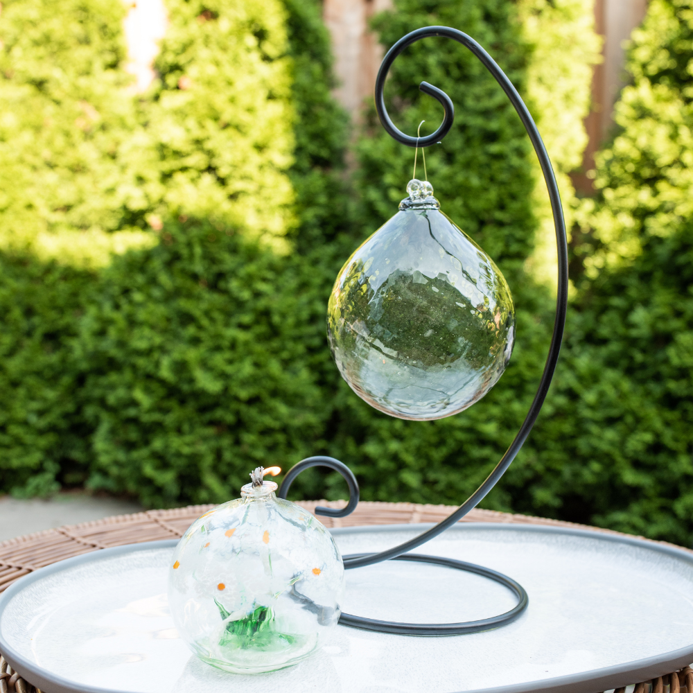 White and green Friendship Blossom Oil Lamp beside a Pewter Enchanting Moonlight Radiance Orb on a Large Single Ornament Holder. Displayed on a white plate in front of green trees