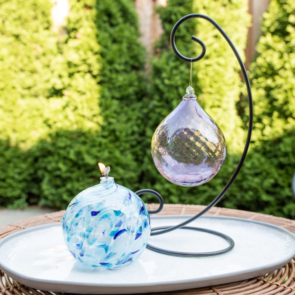 Lit Blue and white Blues Holiday Calico Oil Lamp and Purple Majestic Twilight Radiance Orb on a Large Curved Single Ornament Holder. Displayed on a white plate with green trees in the background