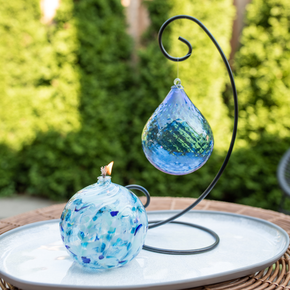 Cobalt Mystical Depths Radiance Orb on a Large Curved Single Ornament Holder. To the left is a lit Blues Holiday Calico Oil Lamp. All on a white place with green trees in the background