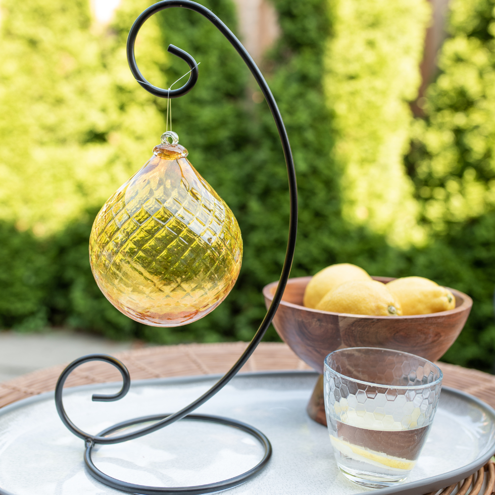 Yellow Sunlit Serenity Radiance Orb on a Curved Single Ornament Holder on a white place. Lemons in a wood bowl and a clear glass of water on the right. Green trees in the background