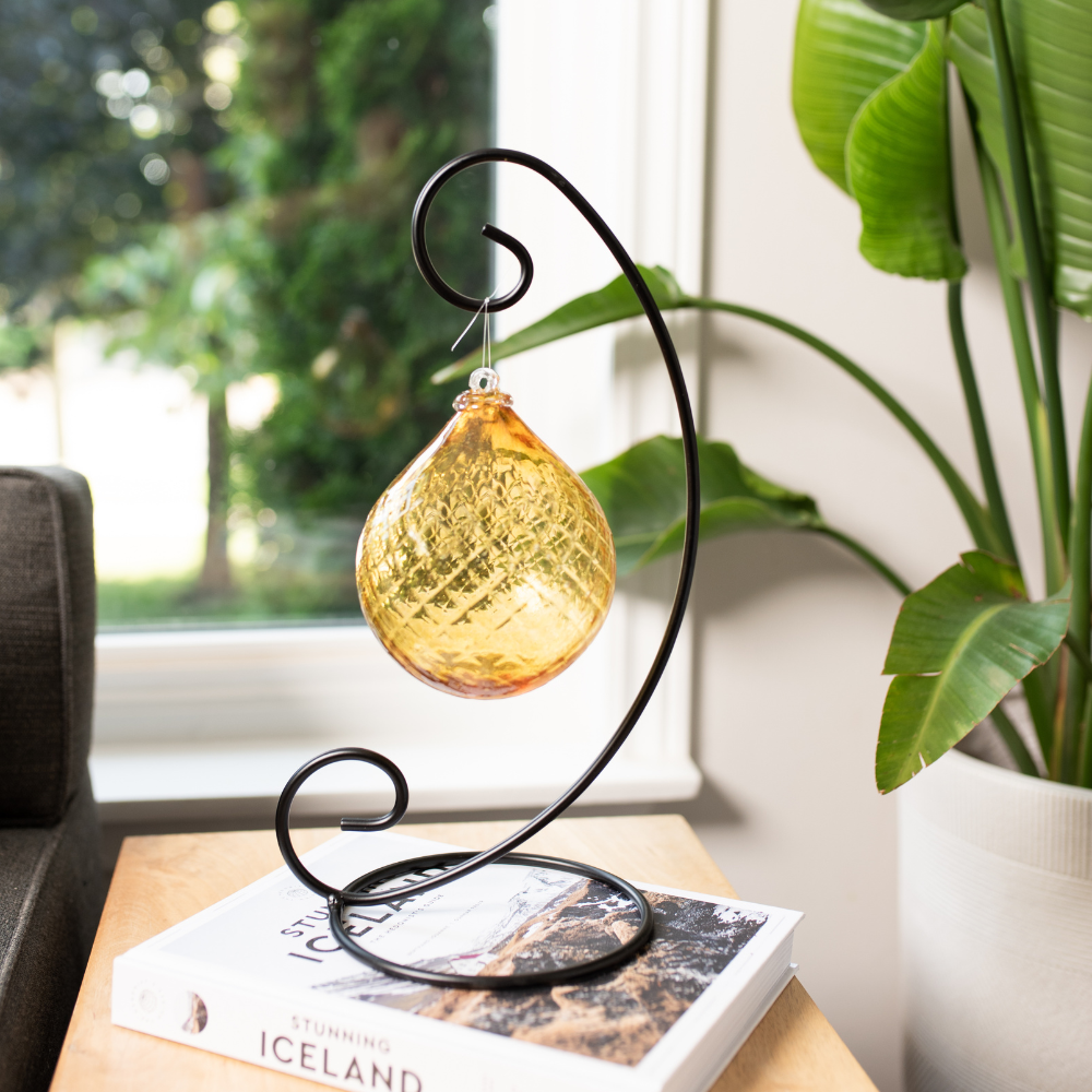 Yellow Sunlit Serenity Radiance Orb on a Large Curved Single Ornament Holder displayed on a white book about Iceland. Window and green plant in the background