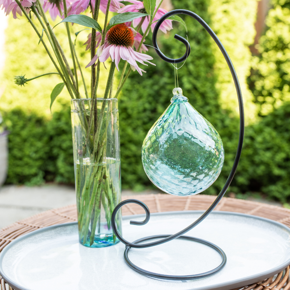 Teal Tranquil Oasis Radiance Orb on a Large Curved Single Ornament Holder with a Teal Friendship Vase full of pink flowers on a white plate with trees in the background