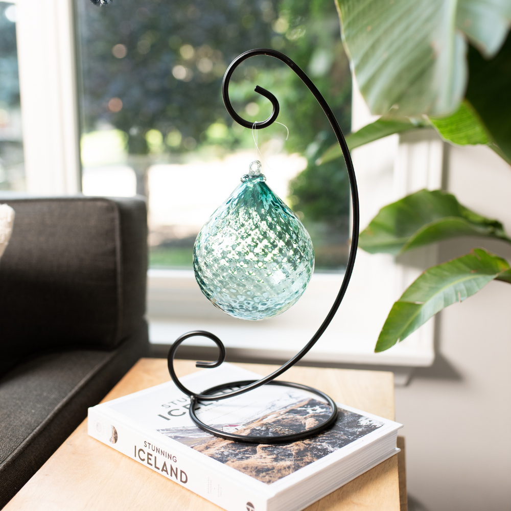 Teal Tranquil Oasis Radiance Orb on a Large Curved Single Ornament Holder resting on a white book with Iceland on the spine. Resting on a wood table. Green plant and window in the background