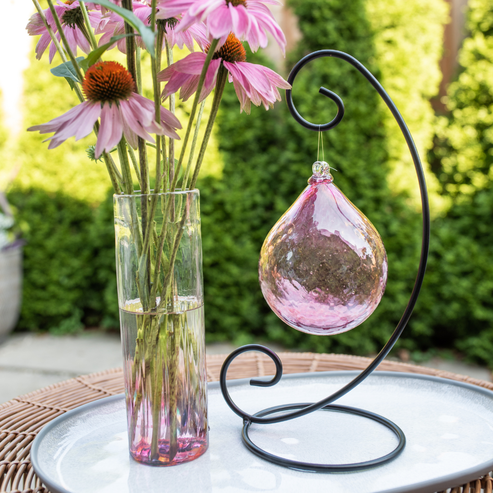 Pink Whispers of Grace Radiance Orb on a Large Curved Single Ornament Holder. Displayed on a white plate beside a pink Friendship Vase. Green trees in the background