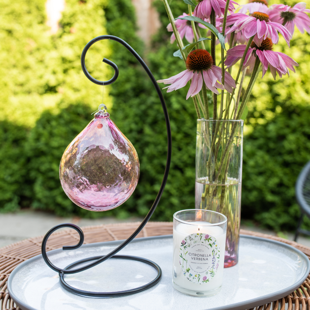 Pink Whispers of Grace Orb on a Large Curved Single Ornament Holder. white candle and Pink Friendship Vase with pink flowers on a white plate. Green trees in the background 