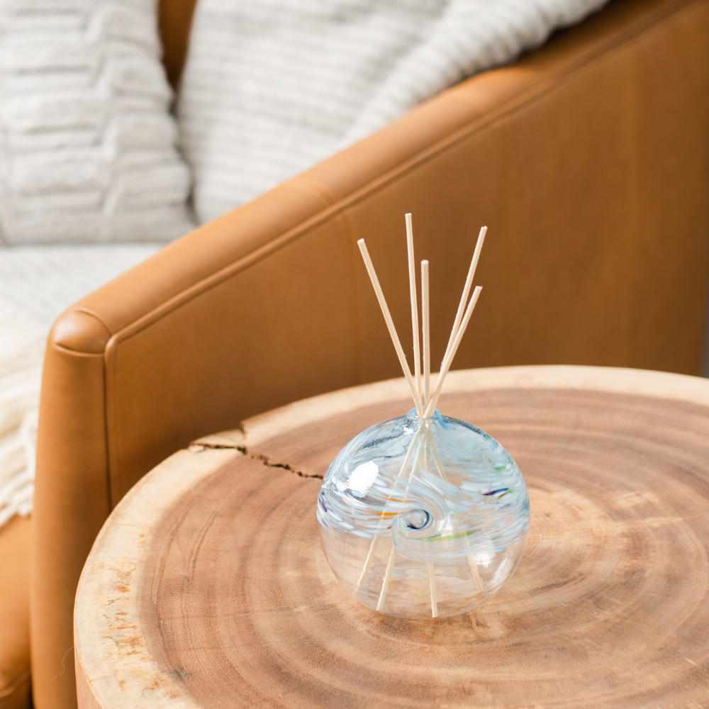 A clear glass orb with a flat bottom with white, blue, red and lime and glass swirled on the top half. Five reeds are in a hole in the top of the orb. Displayed on a wooden table beside a lamp with a book and couch in the background.