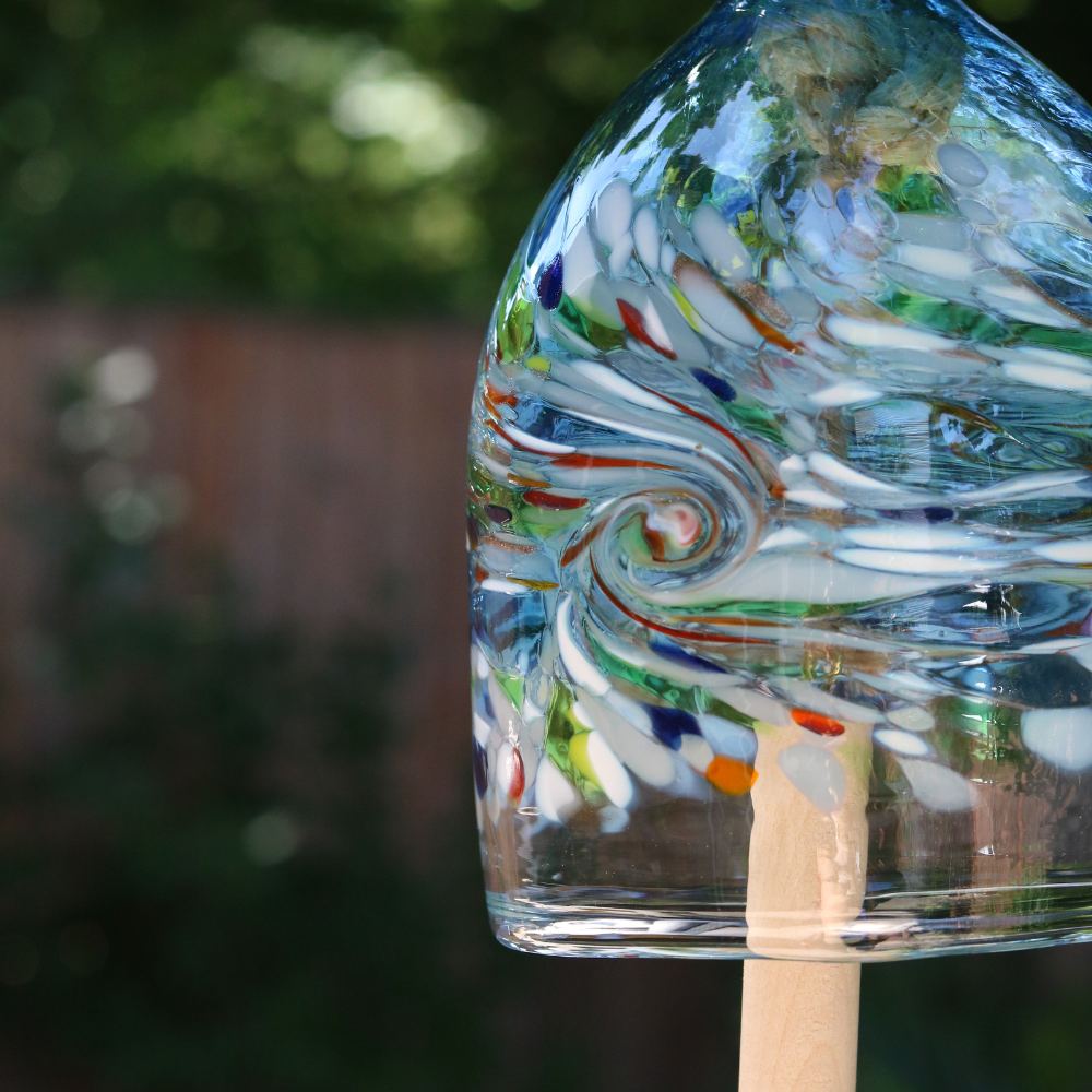 A clear glass bell with opaque white, red, orange, yellow, green and blue glass swirled across the middle. A wooden toggle and rope are pulled through the middle. Hanging amongst greenery.