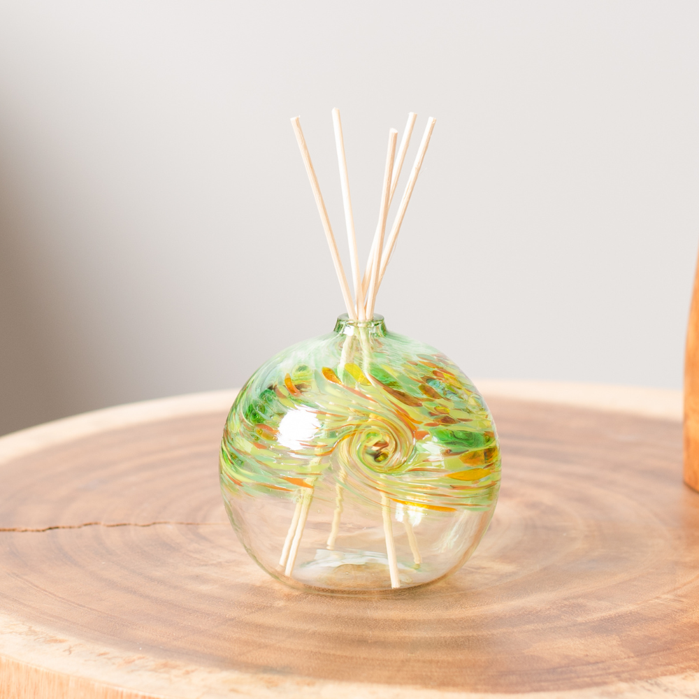 A clear glass orb with a flat bottom with green, amber, red and orange glass swirled on the top half. Five reeds are in a hole in the top of the orb. Displayed on a wooden table. 