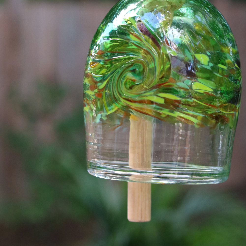 A clear glass bell with green, opaque lime, orange and purple glass swirled across the middle. A wooden toggle and rope are pulled through the middle. Hanging amongst greenery in front of a wood fence.