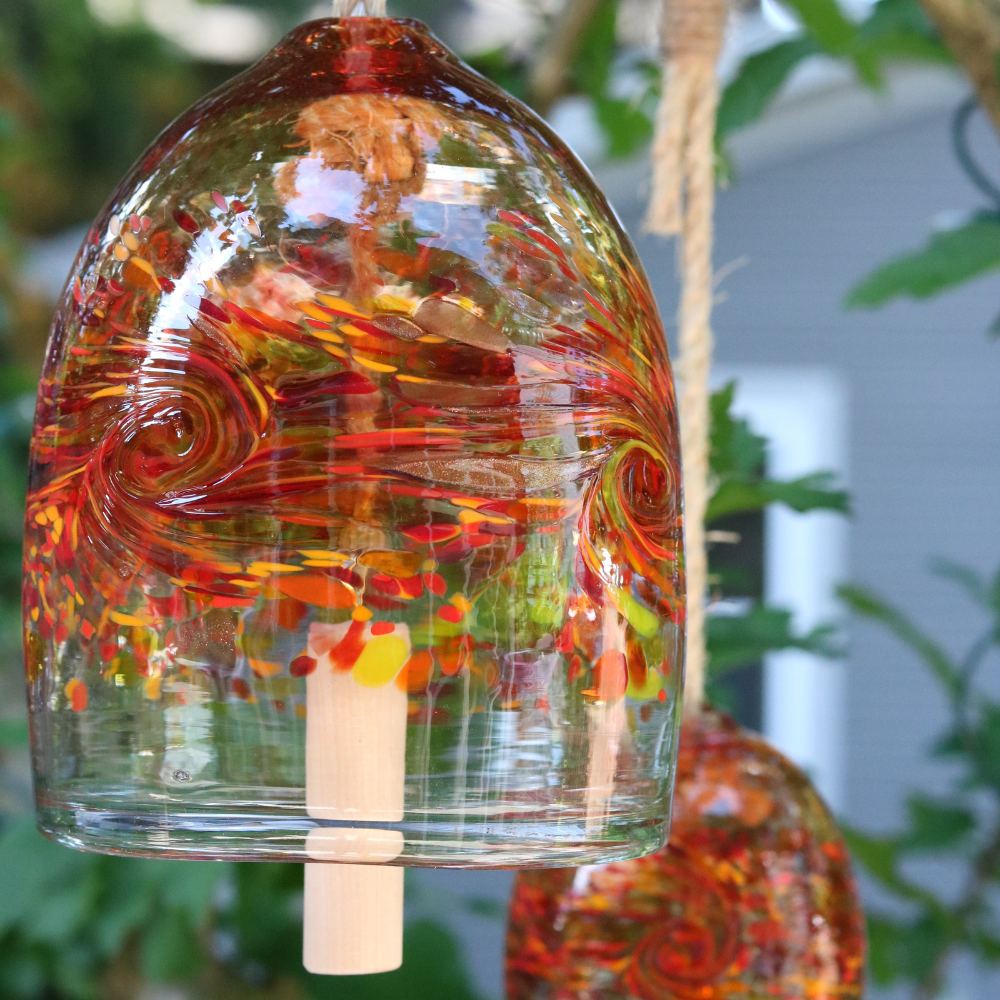 A clear glass bell with red, orange, yellow and purple glass swirled across the middle. A wooden toggle and rope are pulled through the middle. Hanging amongst greenery in front of a wood fence.