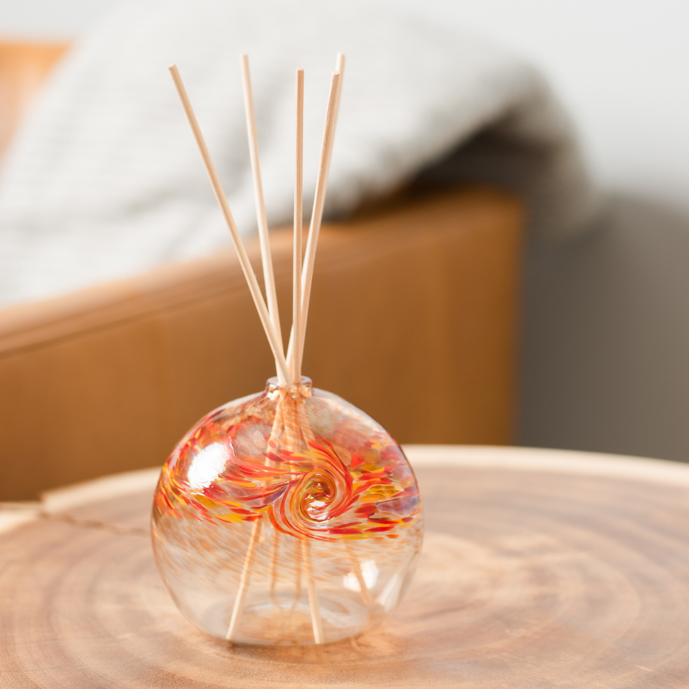 A clear glass orb with a flat bottom with red, orange and yellow glass swirled on the top half. Five reeds are in a hole in the top of the orb. Displayed on a wooden table.