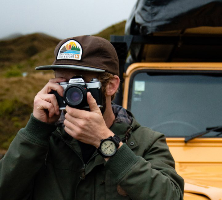 Man holding a camera to his face and wearing a men's analog watch from Nixon stands in front of a jeep on a gloomy day.