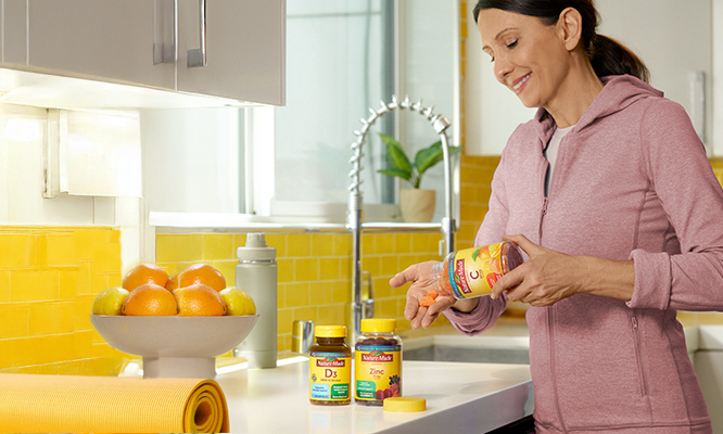 Person holding a bottle of Nature Made Vitamin C immune support supplements, with Vitamin D3 and Zinc bottles and fresh oranges on the counter.