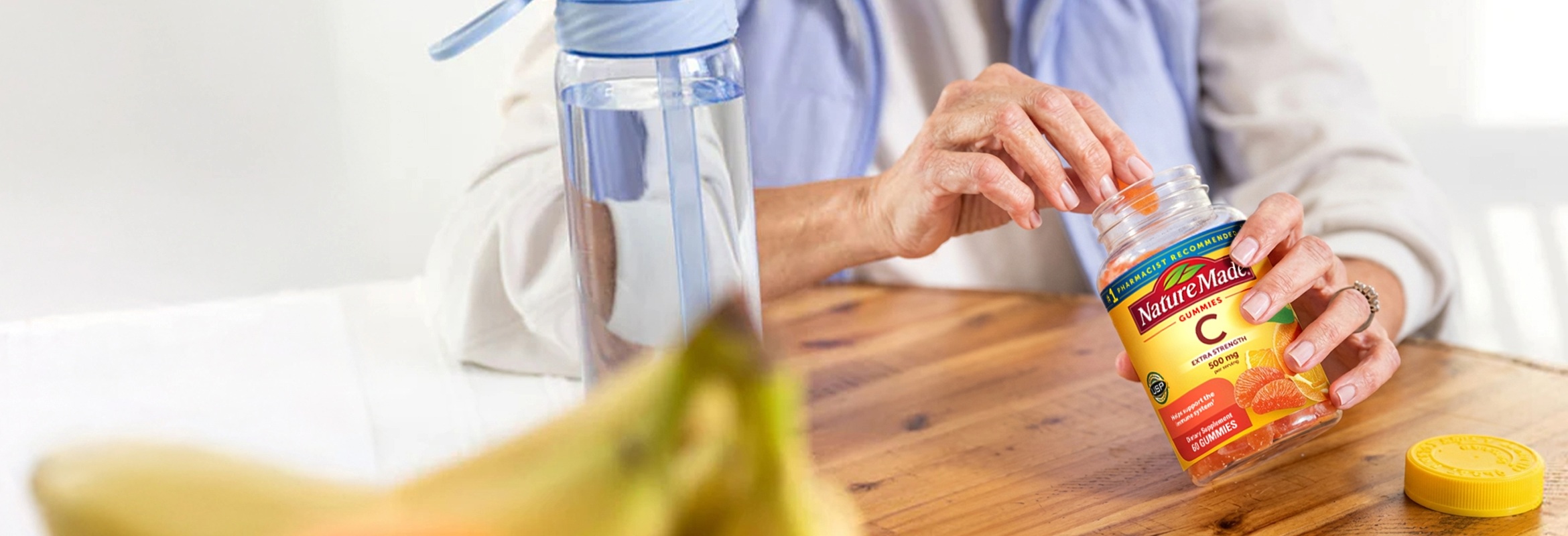 Person reaching for Nature Made Vitamin C gummies at a table with water bottle and fruit.