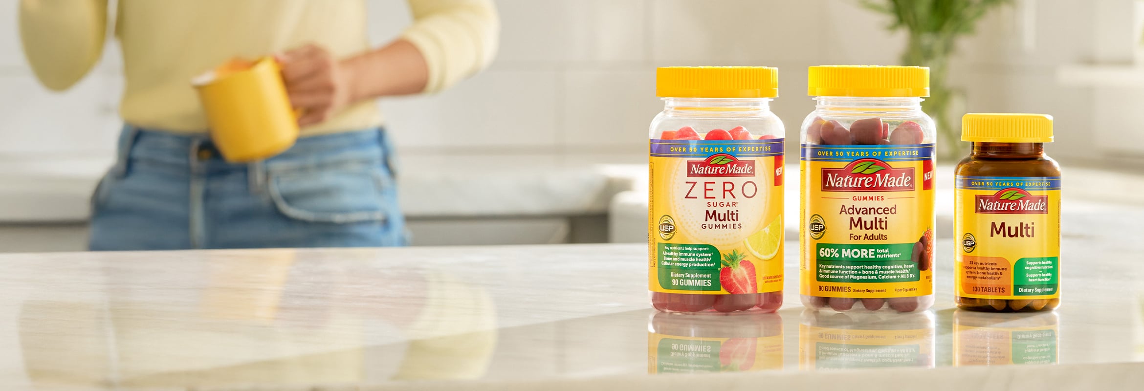 Nature Made Zero Sugar Multi Gummies, Advanced Multi Gummies, and Multi tablets on a kitchen counter with a person holding a yellow mug in the background.