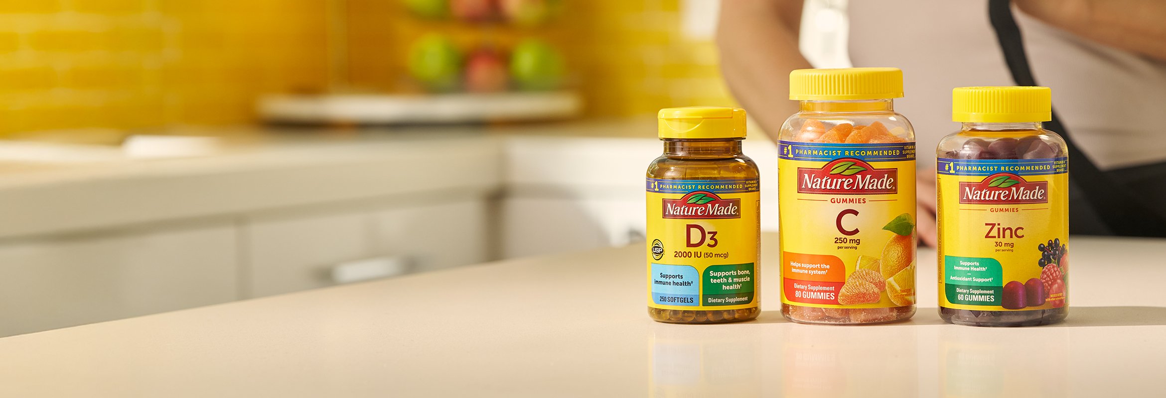 Nature Made immune supplements on a bright kitchen counter with a yellow tile backsplash and a bowl of fresh fruit in the background.