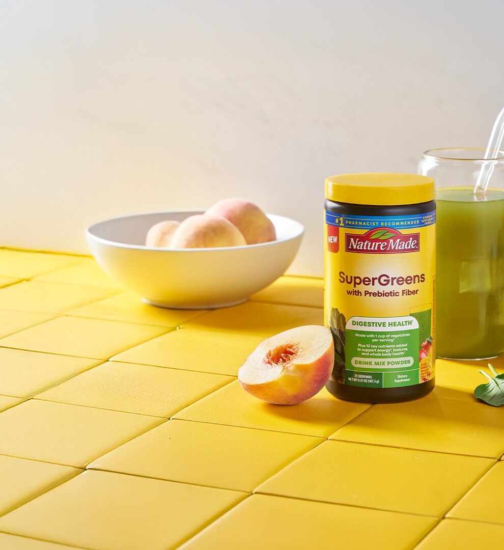 Nature Made SuperGreens With Prebiotic Fiber Mix tub on a counter with a green beverage and fresh fruits and vegetables.