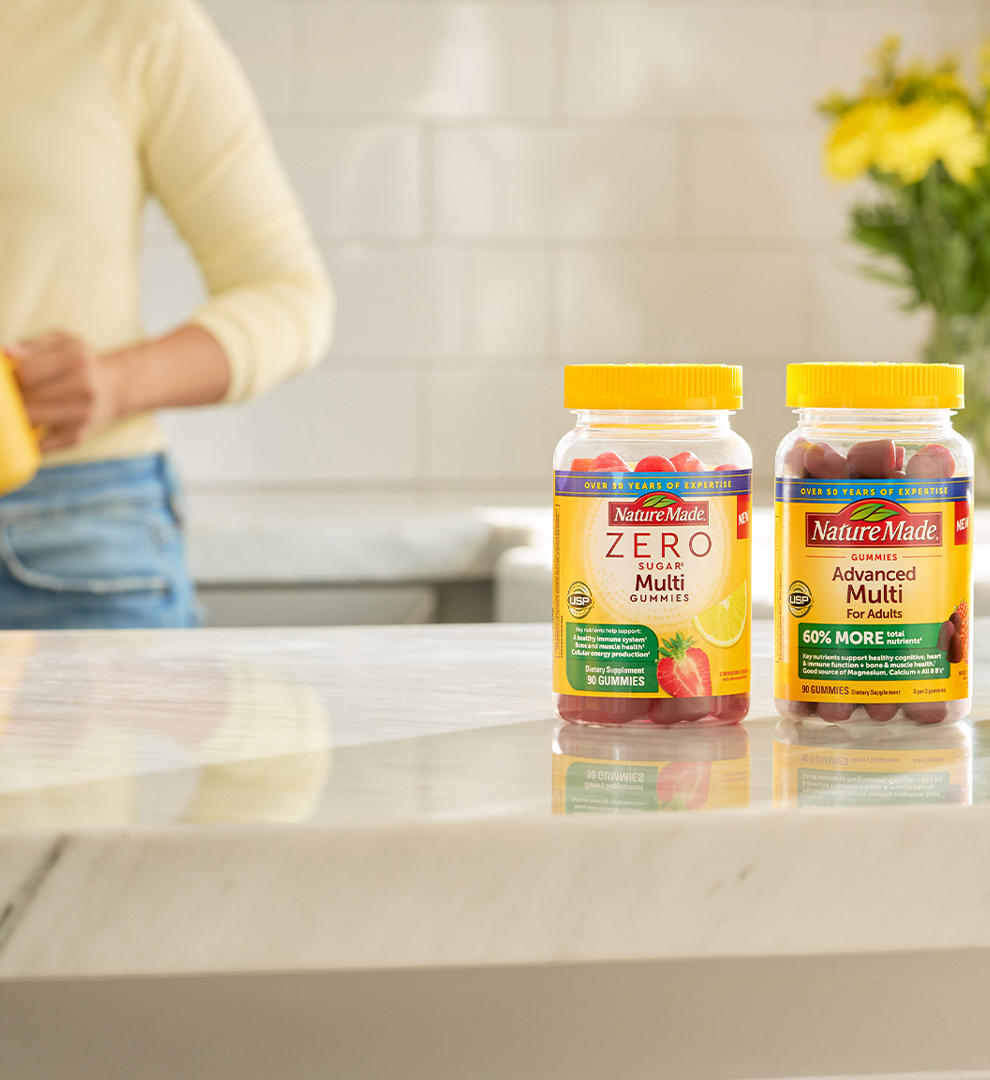 Nature Made Zero Sugar Multi Gummies, Advanced Multi Gummies, and Multi tablets on a kitchen counter with a person holding a yellow mug in the background.