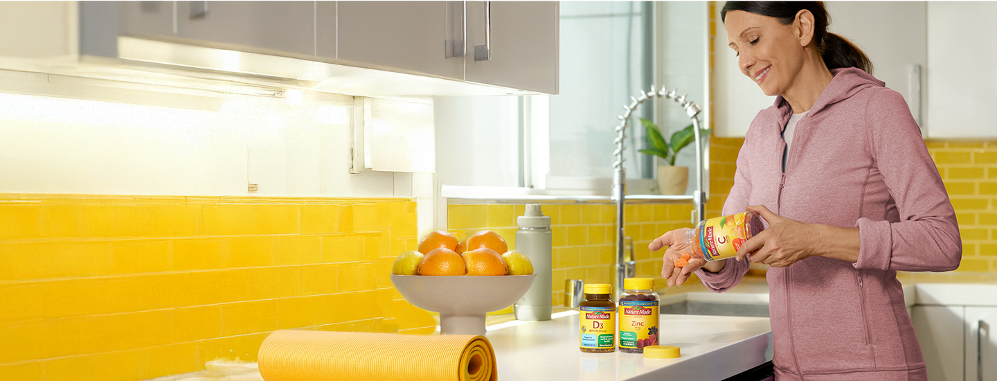 Person holding a bottle of Nature Made Vitamin C immune support supplements, with Vitamin D3 and Zinc bottles and fresh oranges on the counter.