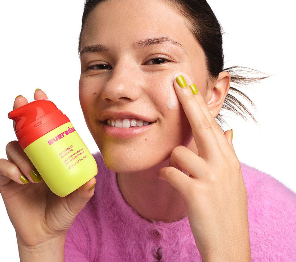 Woman holding Evereden skincare bottle while applying cream to her cheek.